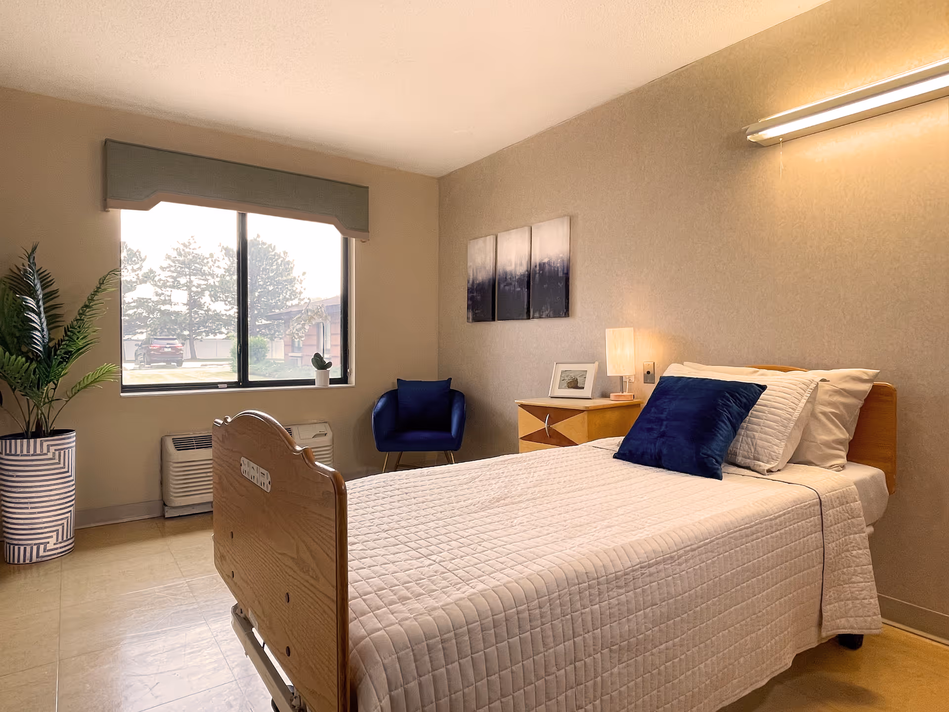 Bright senior-living bedroom with a bed, nightstand, blue chair, plant, and window view.