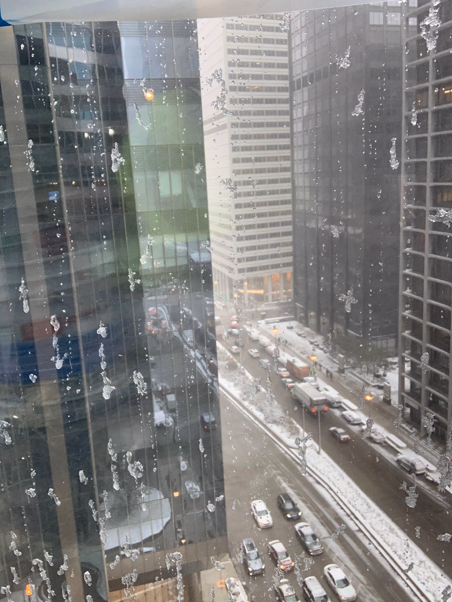 Snow on tall city buildings seen through a rain-splattered window overlooking a snowy street.