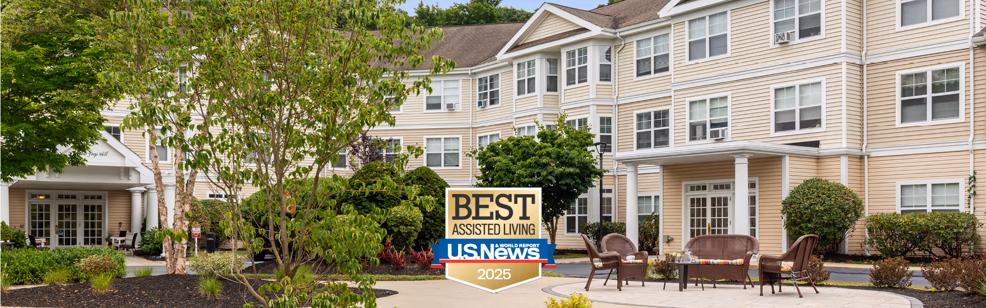 Exterior view of a beige multi-story senior living facility with white trim, surrounded by green trees and shrubs. In front of the building is a sign that reads 'BEST ASSISTED LIVING U.S. News 2025'.