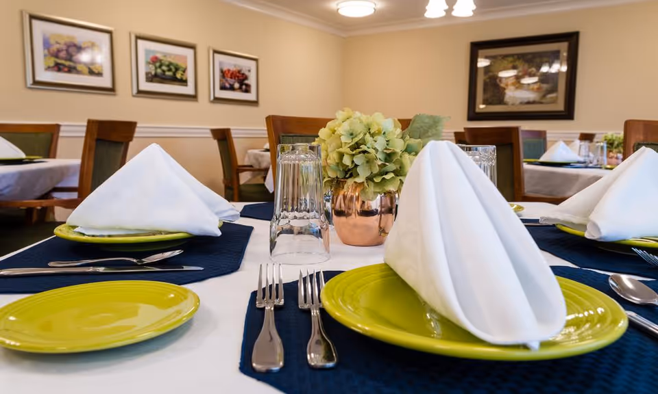 A dining table set with green plates, white folded napkins, silverware, and clear drinking glasses. A small copper vase with green hydrangea flowers is placed in the center. The background shows more tables and chairs, beige walls with framed pictures, and soft lighting.