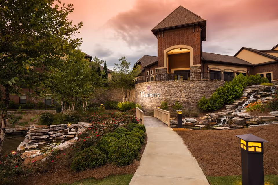Entrance of the Danberry at Inverness building with a stone walkway, landscaped gardens, lanterns, and a cascading pond under a dramatic sky.