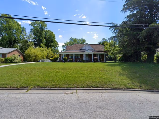 Single-story brick house with a wide front porch, chairs, and a large grassy front lawn under a blue sky.