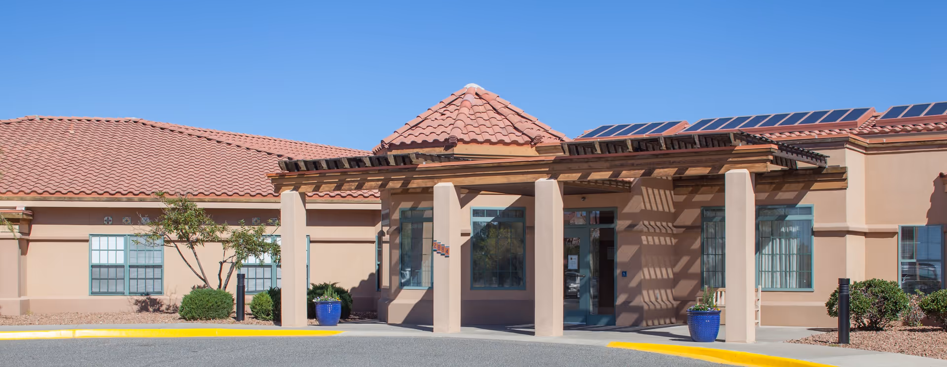 Front exterior view of a senior living facility building with a tiled roof, beige walls, large windows, and a covered entrance supported by columns. There are blue planters with greenery near the entrance and solar panels on the roof under a clear blue sky.