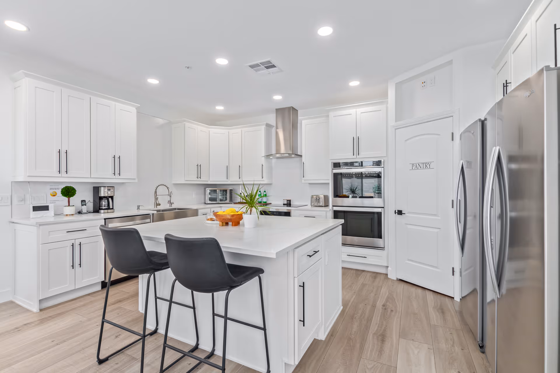 Bright white kitchen with island, stainless appliances, and a labeled pantry in a senior living facility.