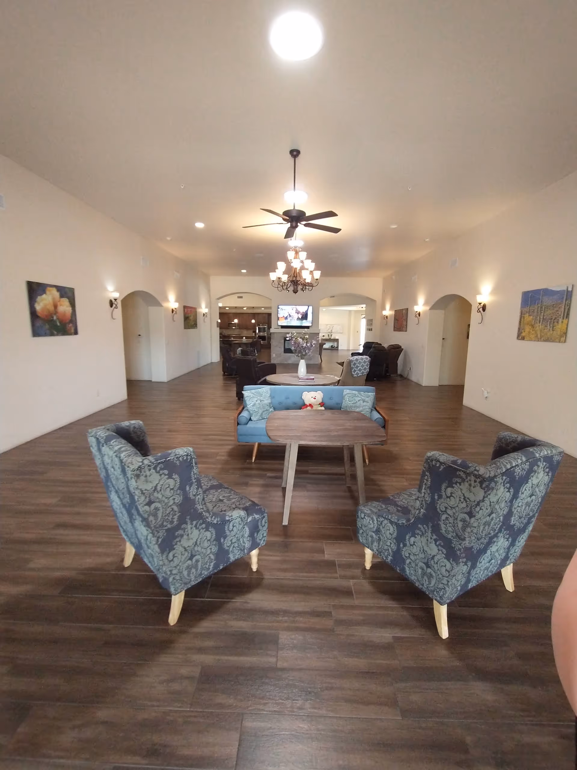 A spacious assisted living lounge with blue patterned chairs around a central wooden table.