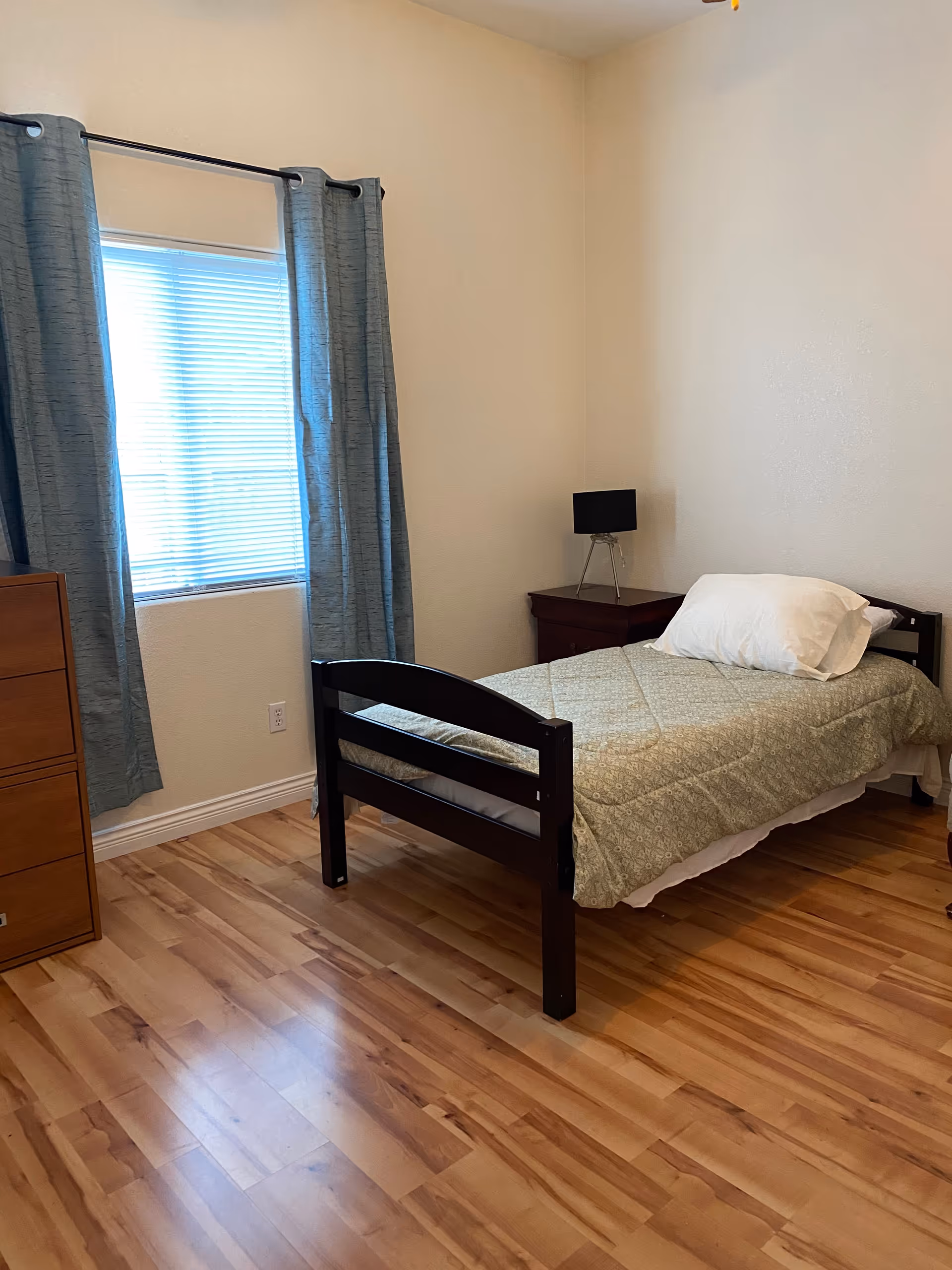 A small bedroom with a single bed, dresser, nightstand, and window with curtains.