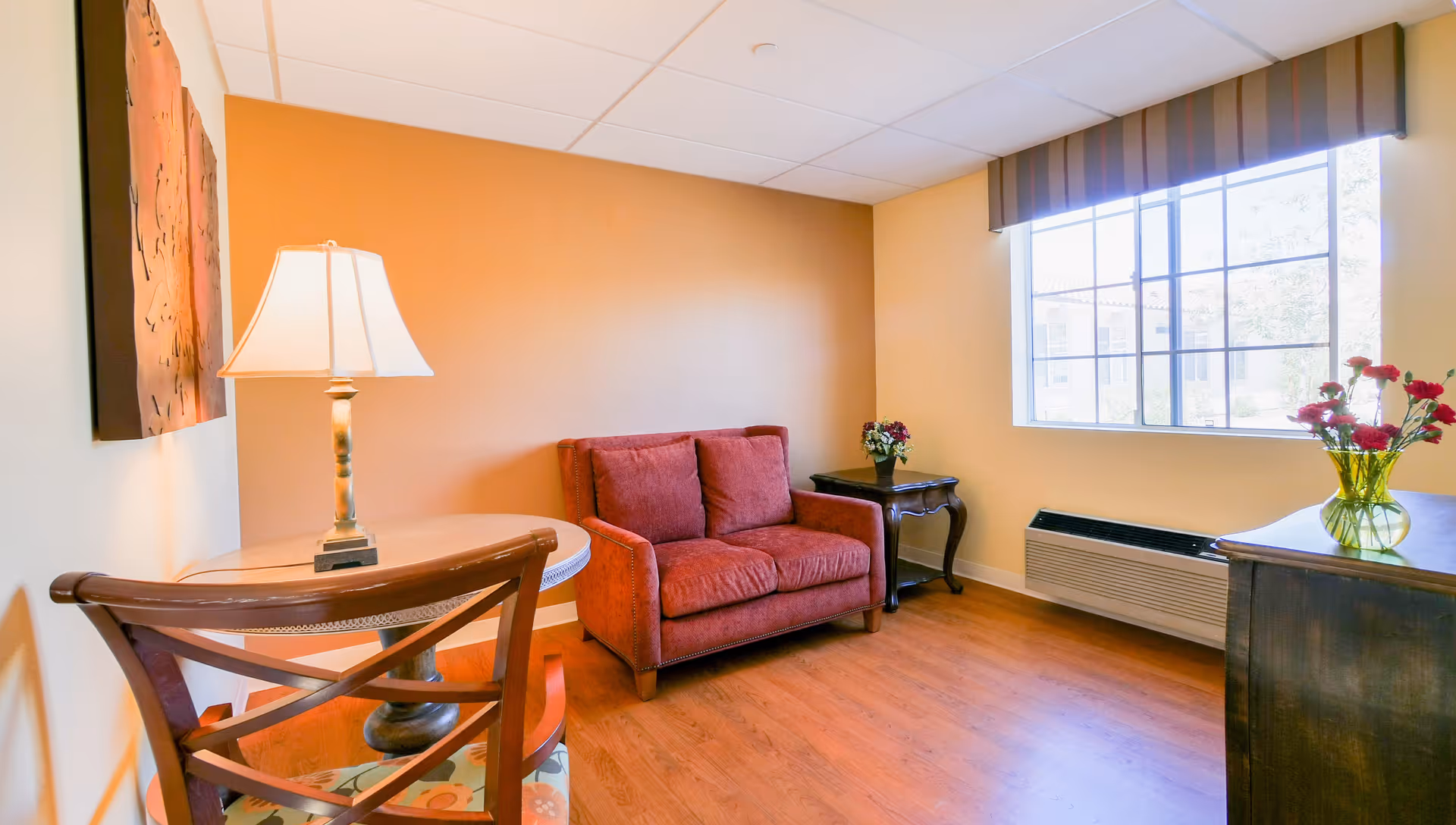 A cozy sitting area in a senior living facility with a red loveseat, a wooden chair and round table with a lamp, a side table with a small flower arrangement, and a large window with striped valance letting in natural light. The walls are painted in warm tones and the floor is wooden.