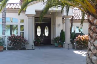 Entrance of a building with double oval glass doors, flanked by columns and surrounded by potted plants and flowers. Palm trees are visible in the foreground and the building has a tiled roof.