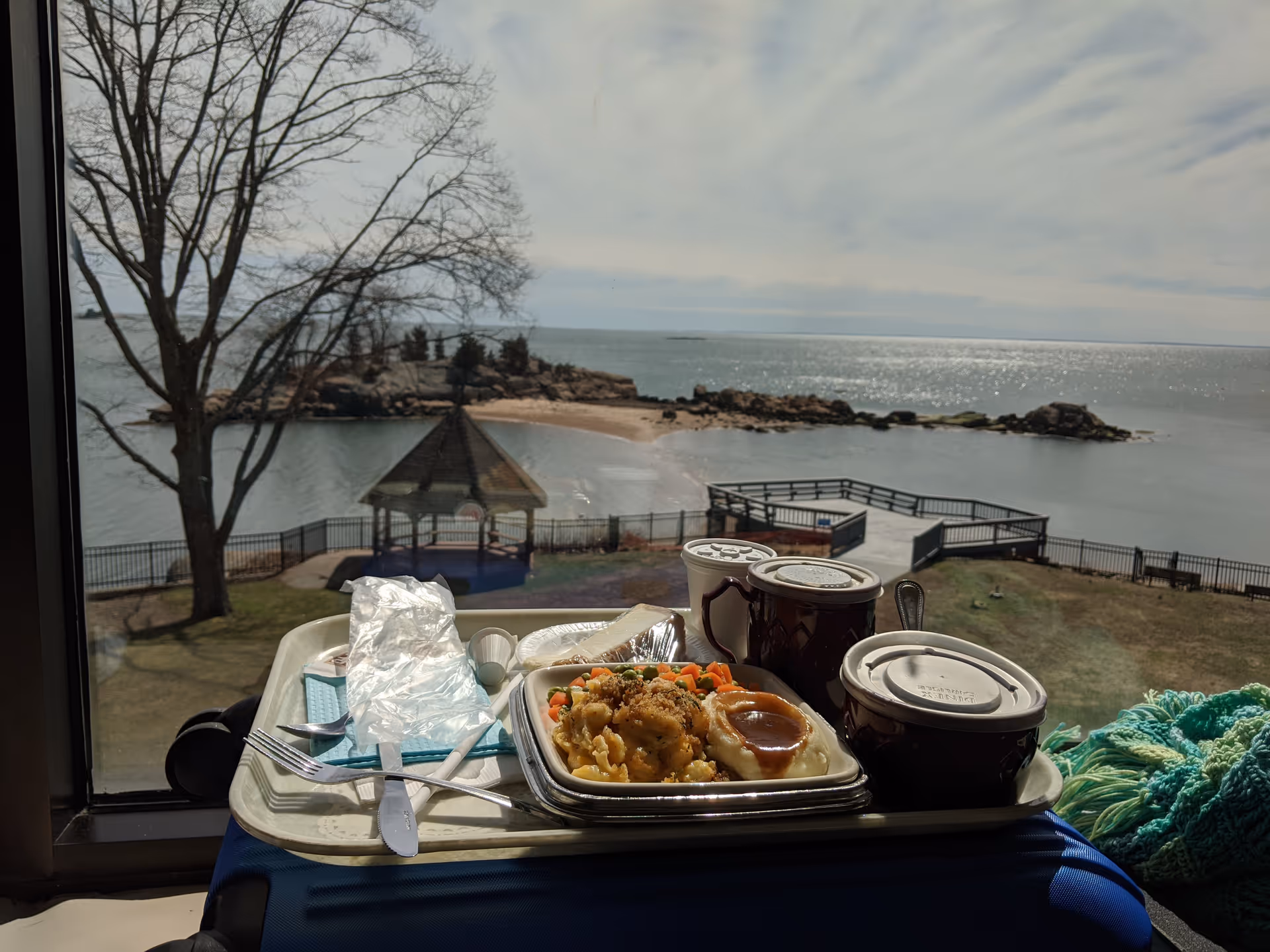 A meal tray with utensils and takeout dishes on a windowsill overlooking the sea.
