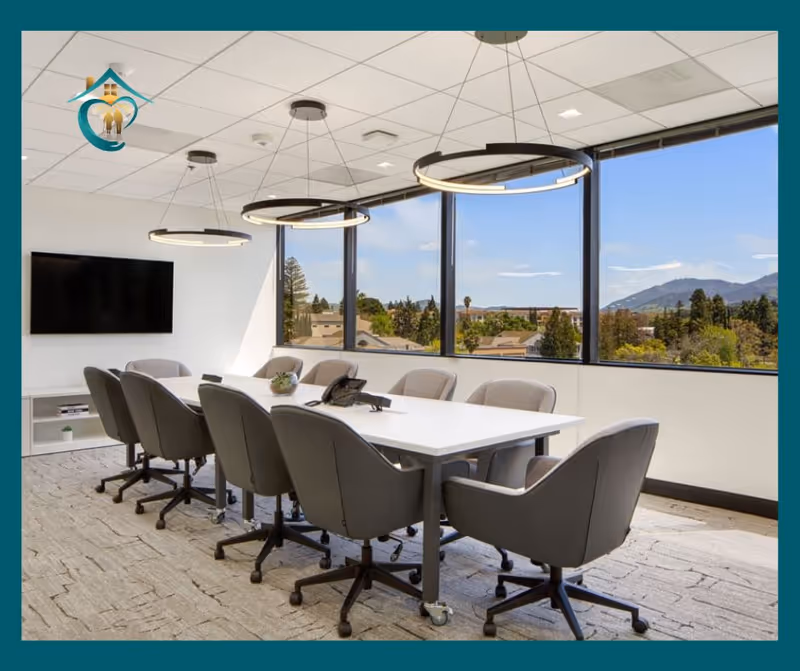 Bright conference room with a long white table, grey chairs, and large windows overlooking landscape.
