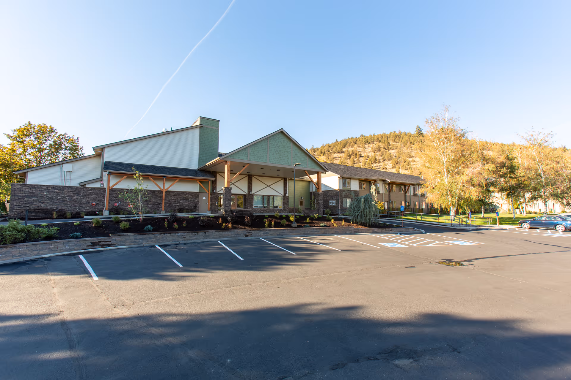 Front exterior of a retirement community building with a covered entrance, empty parking spaces, and a hillside in the background.