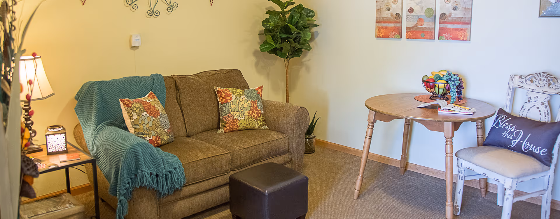 A cozy living room area with a brown sofa adorned with two floral patterned pillows and a teal throw blanket. Next to the sofa is a small side table with a lamp and decorative items. A small dark ottoman is placed in front of the sofa. On the right side, there is a round wooden table with a bowl of fruit and an open book, accompanied by a white distressed chair with a cushion and a pillow that says 'Bless this House'. A tall green plant is positioned in the corner, and two colorful framed artworks hang on the wall above the table.
