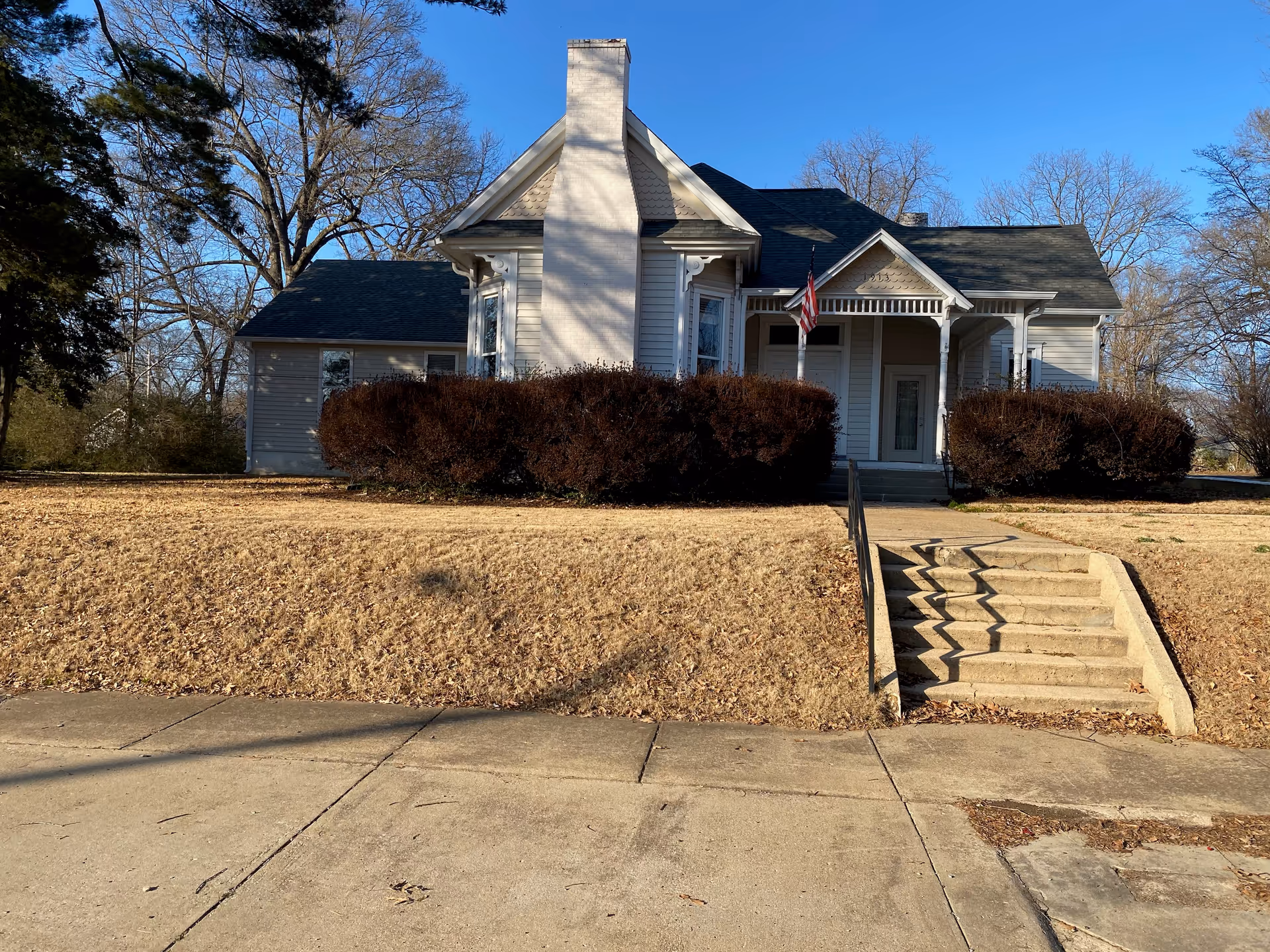 Front view of a white house with steps, shrubs, and a flag by the entrance.