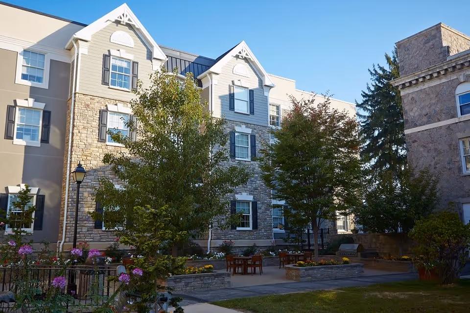 Courtyard with patio tables, trees, and flower beds in front of a multi-story stone-and-siding senior living building.