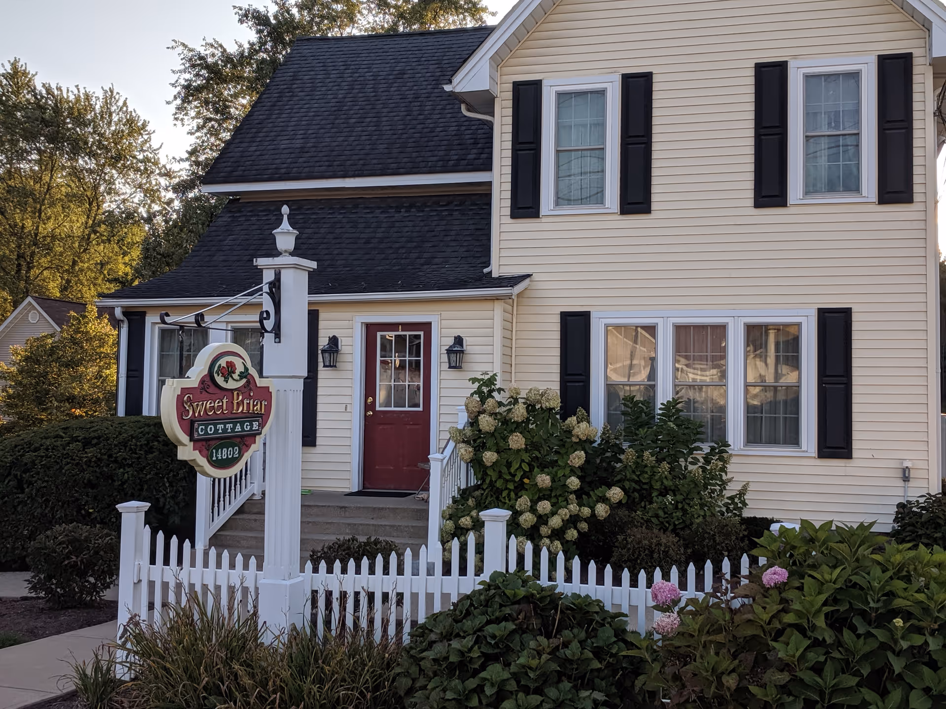 Two-story beige cottage with white picket fence, hydrangea shrubs, and a Sweet Briar Cottage sign.