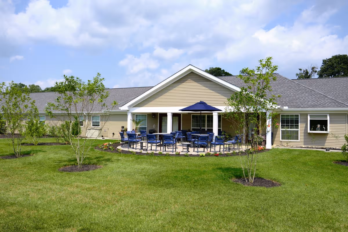 Outdoor patio area of a senior living facility with multiple blue chairs and tables arranged under a blue umbrella, surrounded by green grass and small trees, with a beige building in the background under a partly cloudy sky.