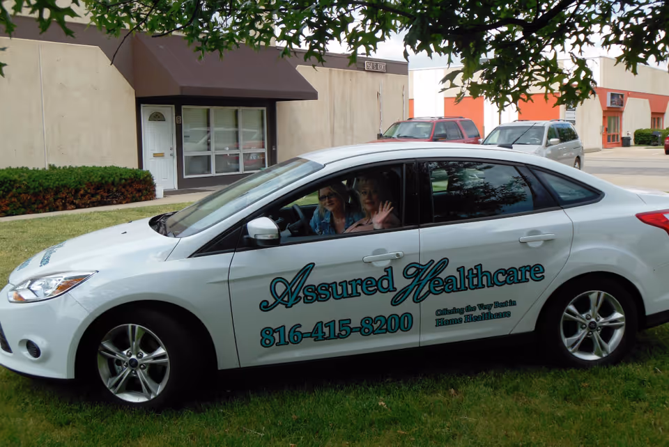 A white Assured Healthcare car parked on grass in front of a building.