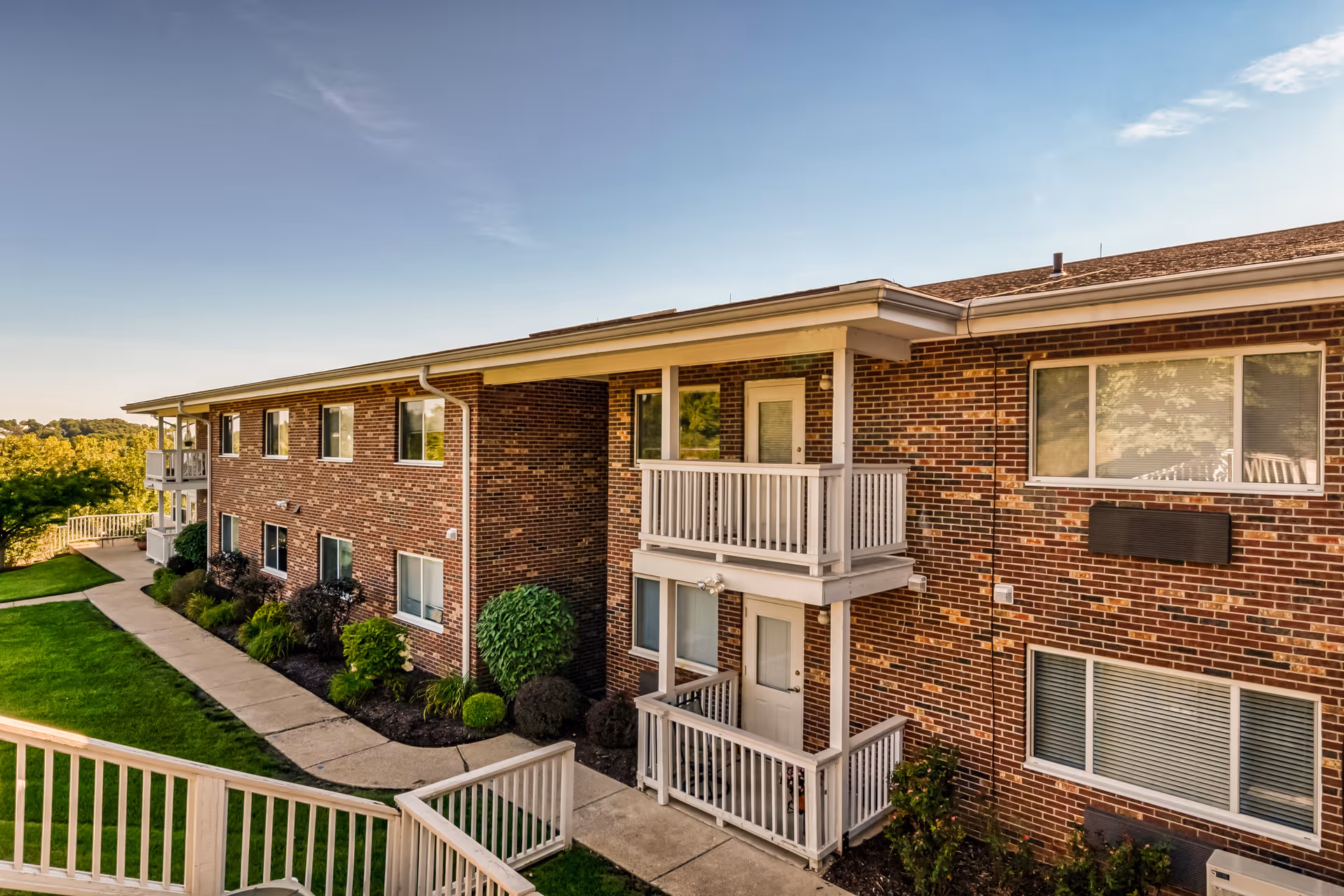 Exterior view of a two-story brick building with white balconies and railings, surrounded by green grass and landscaped bushes under a clear blue sky.