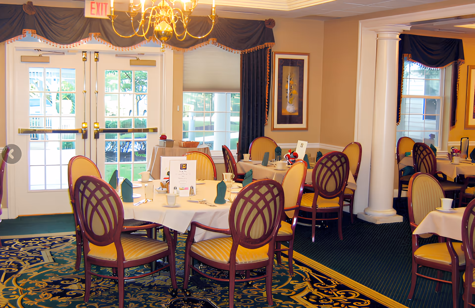 A dining room with round tables covered in white tablecloths, set with cups, napkins, and menus. The room features elegant wooden chairs with yellow upholstery, large windows with dark curtains, a chandelier, and decorative columns.