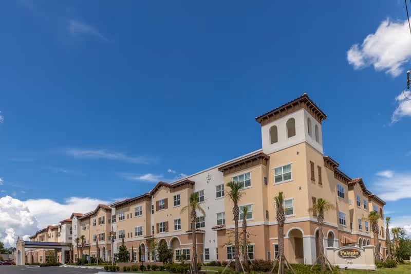 Exterior view of a beige multi-story senior living building with palm trees and blue sky.
