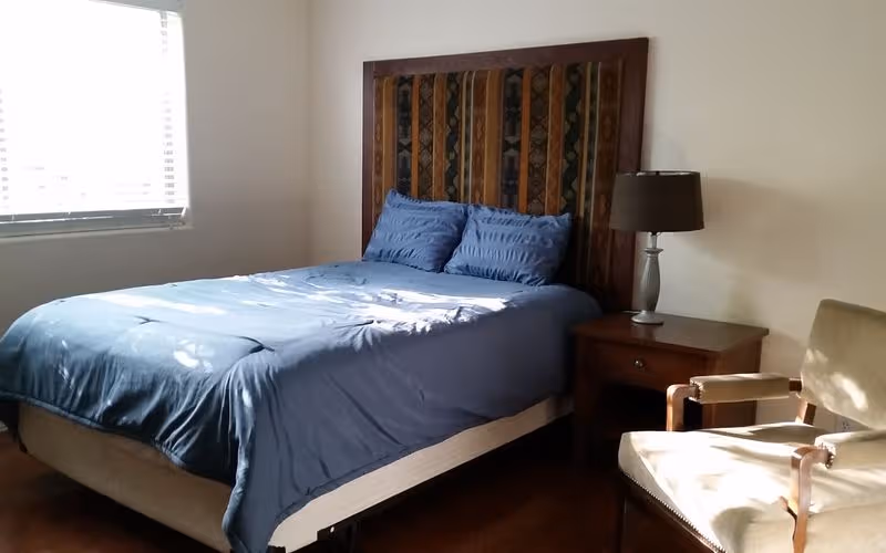 A neatly made blue comforter on a bedroom bed with a nightstand, lamp, and chair.