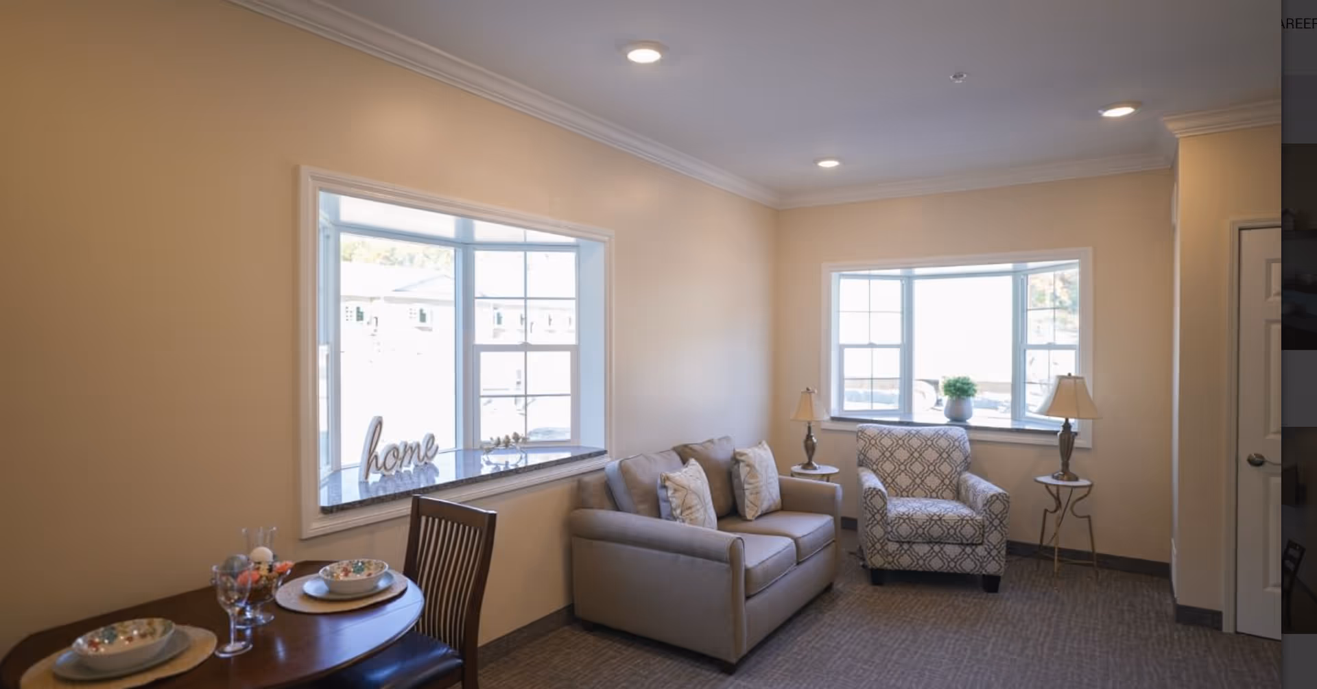 A cozy living room area with a beige sofa adorned with three pillows, a patterned armchair, two small side tables each with a lamp, and two large windows letting in natural light. A small dining table with two place settings is partially visible in the foreground. The walls are painted a soft beige color and the carpet is a neutral tone.