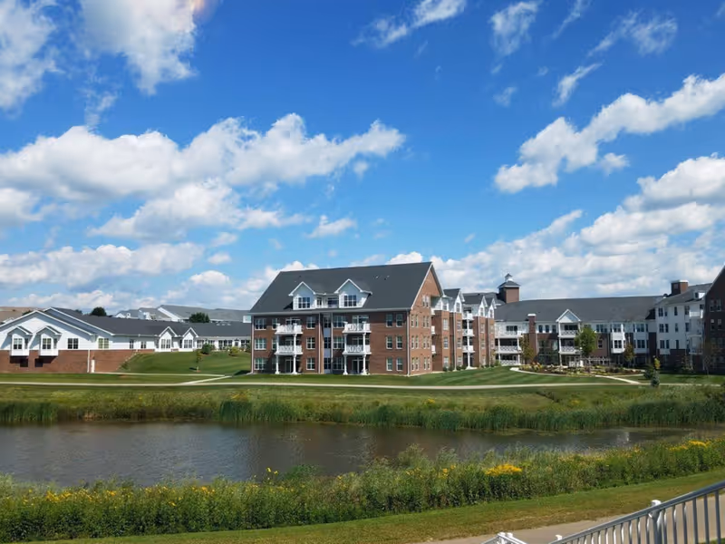 A large senior living facility with multiple connected buildings featuring brick and white siding exteriors, set against a bright blue sky with scattered clouds. In the foreground, there is a pond surrounded by green grass and wildflowers.