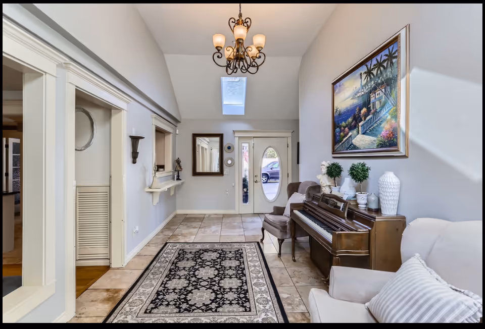 Bright foyer with piano, chandelier, seating, and tiled floor in a senior living lobby.