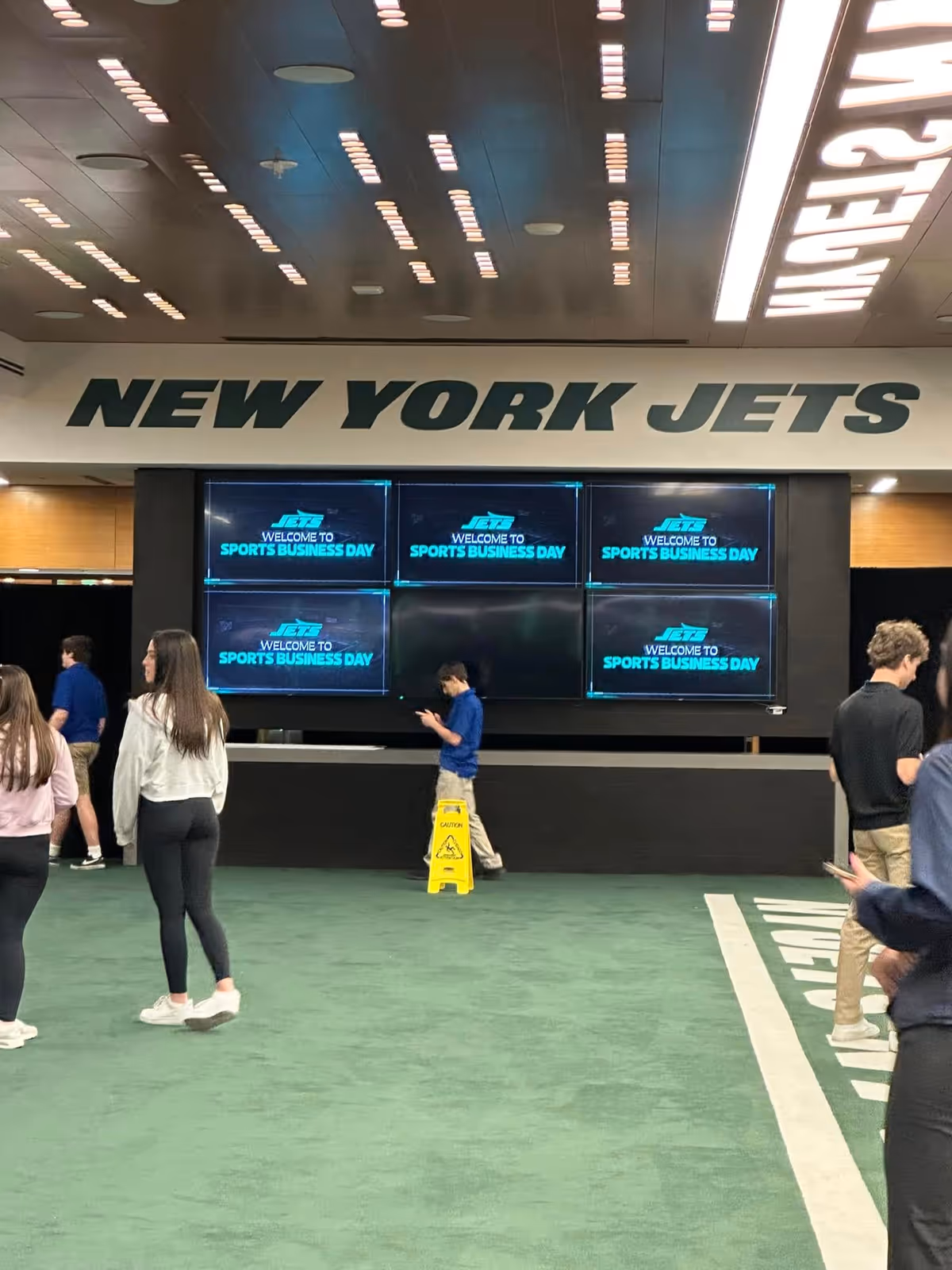 Indoor sports venue with New York Jets signage, multiple screens, and people walking on green floor.
