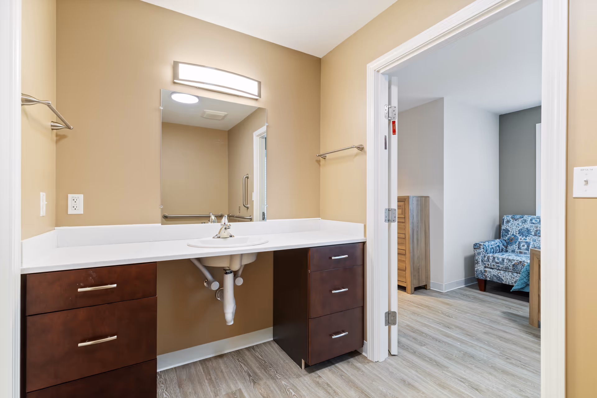 Accessible bathroom vanity with countertop sink, mirror and light, and an open door revealing a bedroom with a patterned armchair.