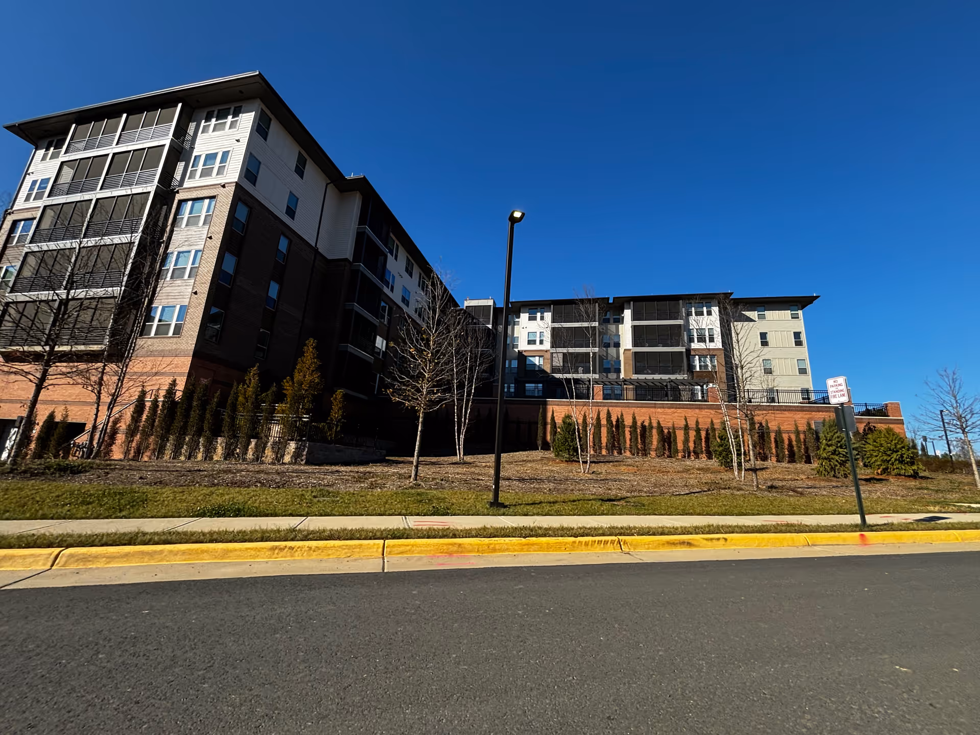 Exterior view of a multi-story senior living facility building under a clear blue sky, with landscaped grounds including small trees and shrubs, a sidewalk, and a street with a yellow curb and a no parking sign.