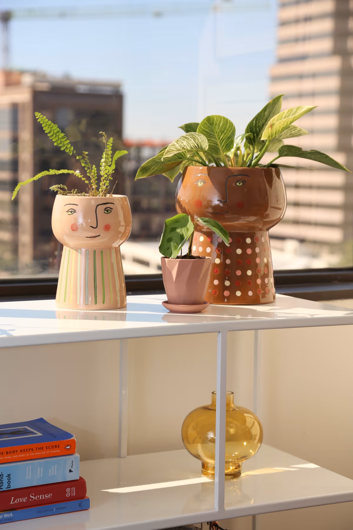 Sunlit interior shelf with two face pots and a small yellow vase by a window.
