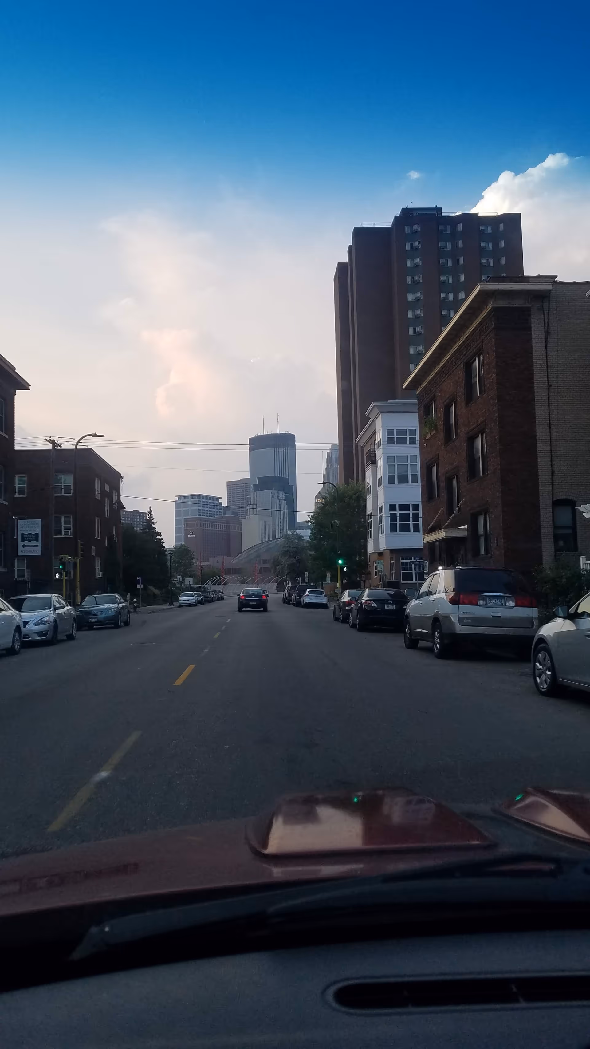 Street view of a city avenue with parked cars and tall buildings under a blue sky.