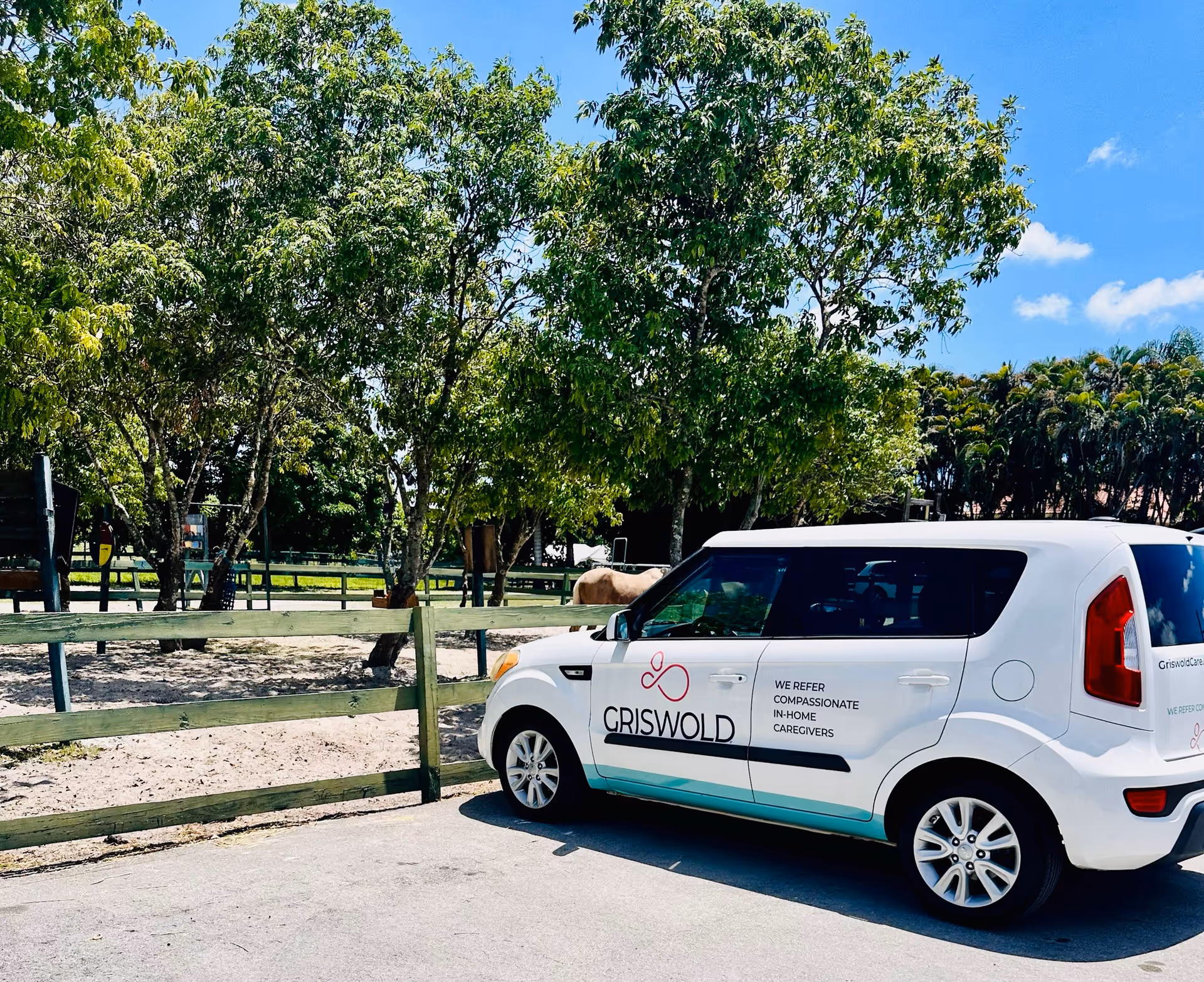 White Griswold van parked beside a wooden fence under green trees in a sunny outdoor area.