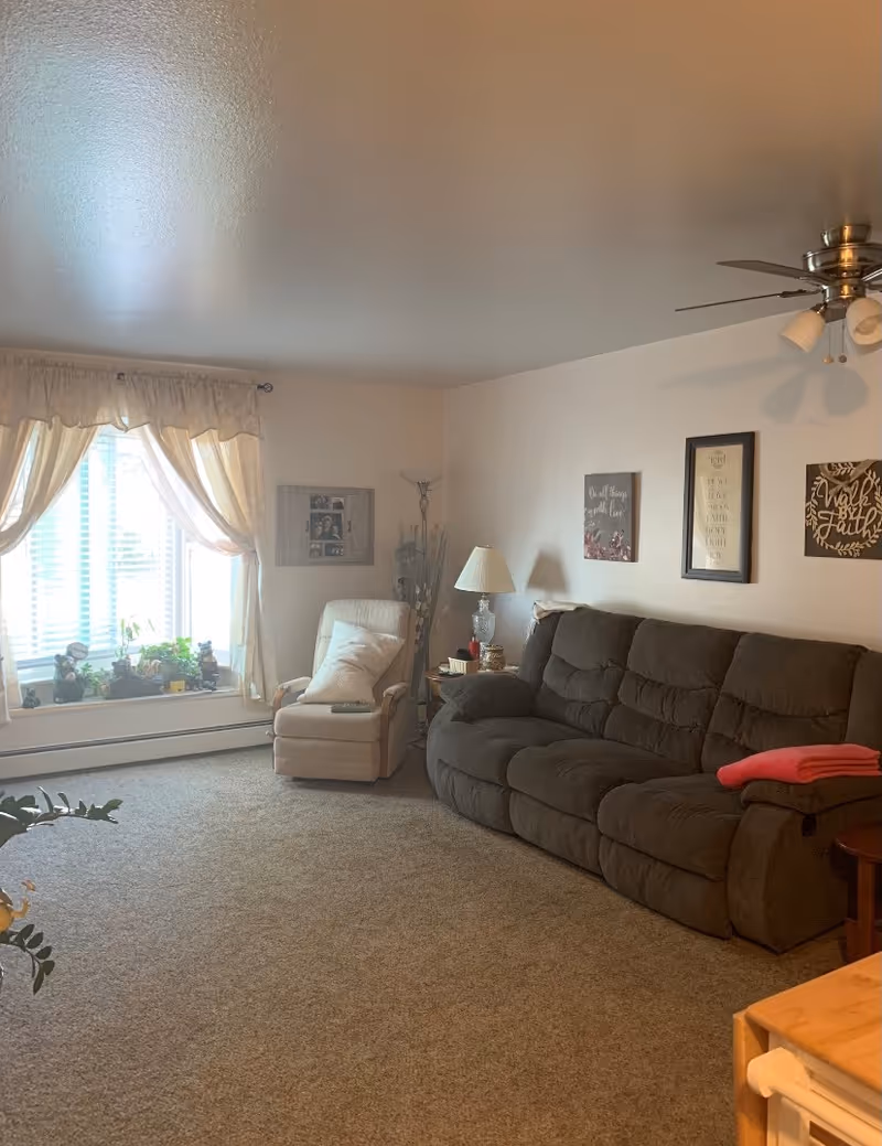 Cozy living room with a large dark reclining sofa, an armchair by a curtained window, side table with a lamp, wall art, and carpeted floor.