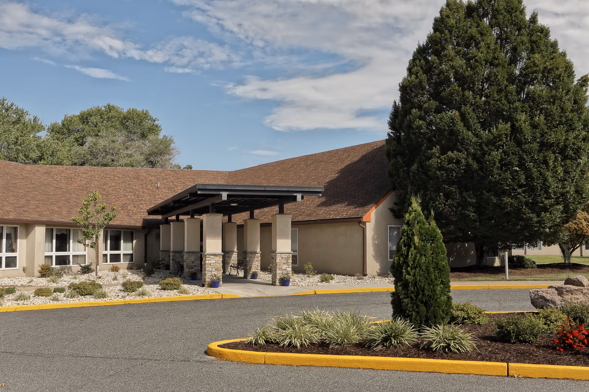Exterior view of a single-story building with a brown shingled roof and beige walls, featuring a covered entrance supported by stone pillars. The surrounding area includes landscaped greenery, bushes, trees, and a curved driveway with a yellow curb.