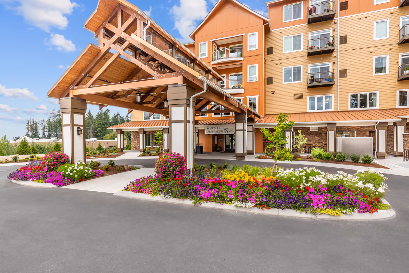 Entrance of Wesley Bradley Park senior living facility with a large wooden canopy, colorful flower beds, and a multi-story building in the background under a partly cloudy sky.