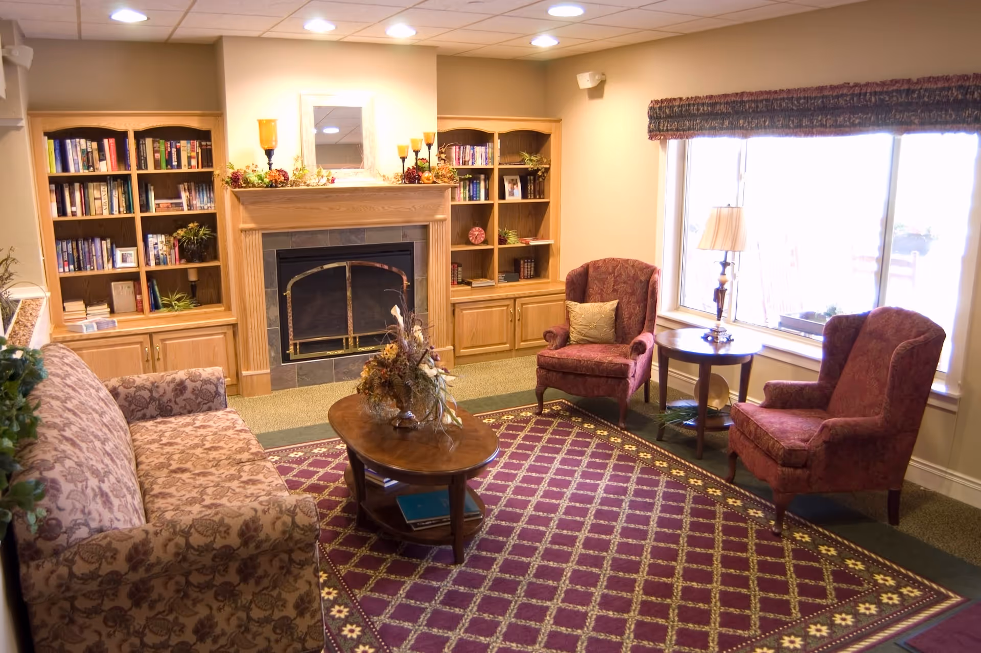 Cozy lounge with fireplace, bookshelves, and seating area by large window.