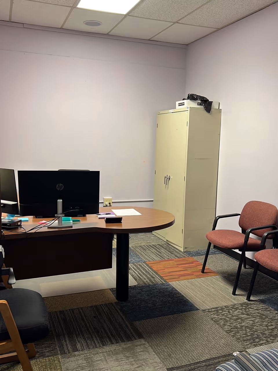 Office desk with dual computer monitors, cabinet, and chairs in a small administrative room.