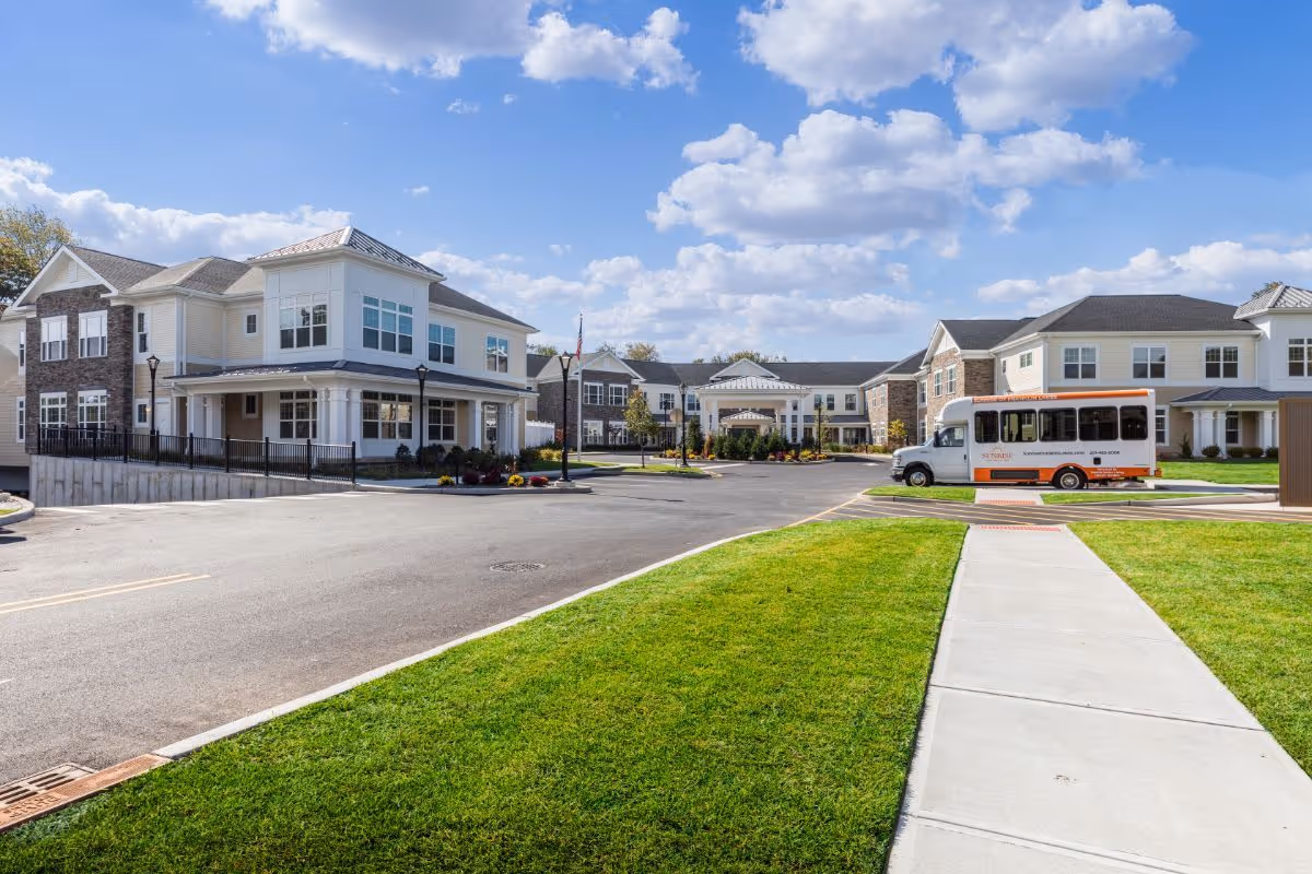 Exterior front of Sunrise of Franklin Lakes senior living campus showing two-story buildings, a shuttle bus, driveway, sidewalk and lawn under a blue sky.