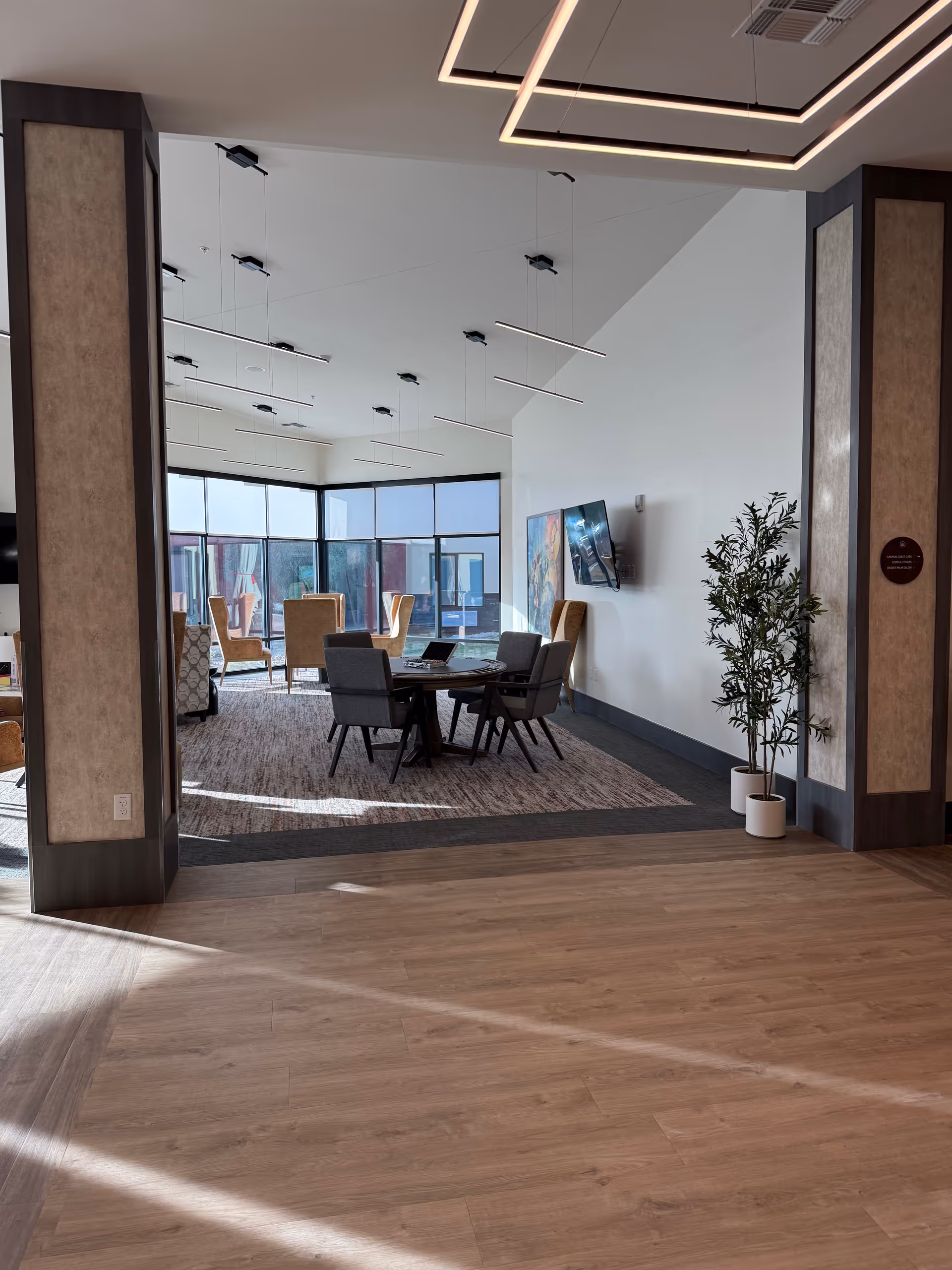 Modern senior living common area with a round table, chairs, TV, and large windows.