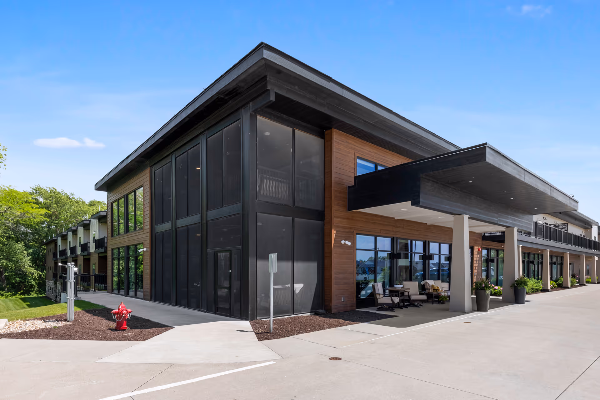 Exterior view of a modern senior living facility building with large windows, a covered entrance with seating, and a clear blue sky.