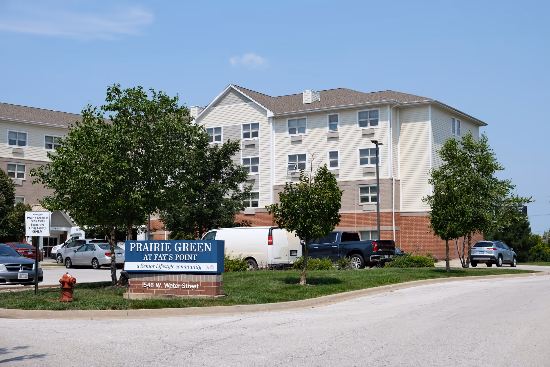 Exterior view of Prairie Green at Fay's Point senior living facility showing a multi-story building with beige and brick facade, several trees, parked cars, and a blue sign with the facility name and address at 1546 W. Water Street.