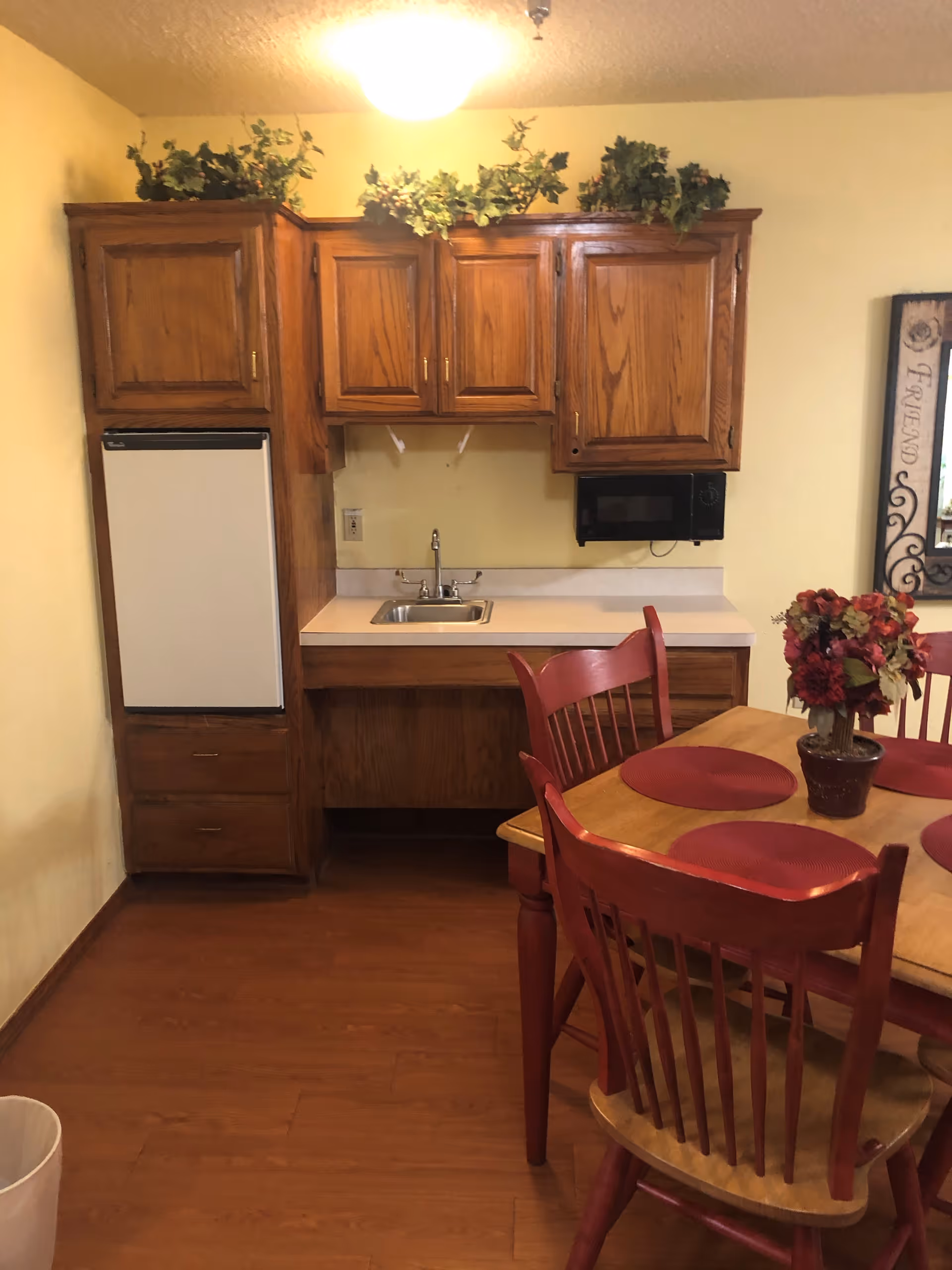 A small kitchen area with wooden cabinets, a white mini refrigerator, a stainless steel sink, and a black microwave mounted under the cabinets. There is a wooden dining table with red chairs and red placemats, and a flower arrangement in a pot on the table. The walls are painted yellow and there is a decorative framed mirror on the right side.