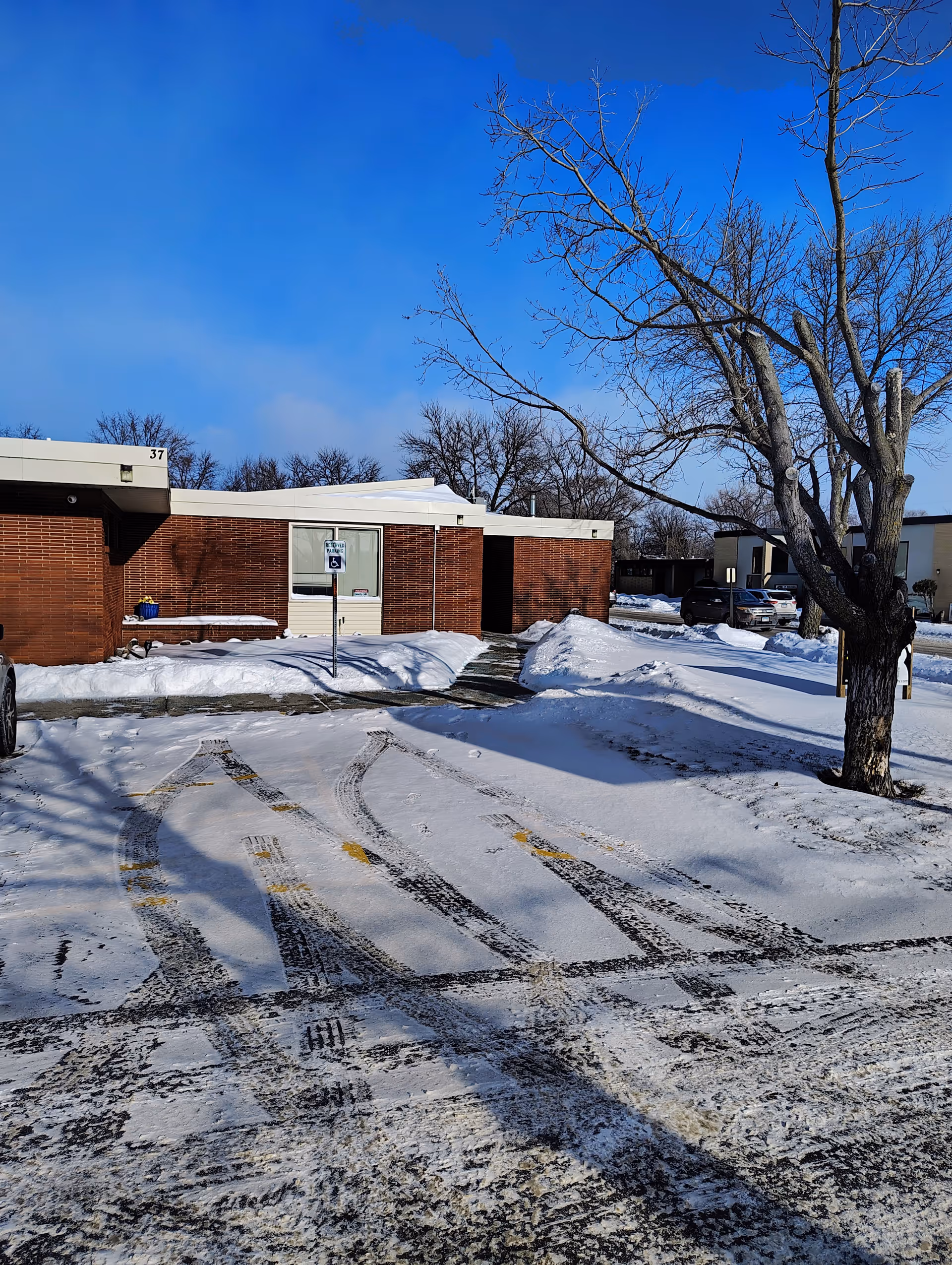 Snow-covered parking lot and brick building exterior with a tree in foreground.