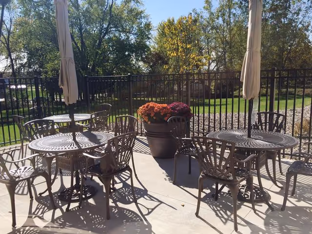 Outdoor patio area with metal tables and chairs, two closed umbrellas, and a large planter filled with colorful flowers. The patio is enclosed by a black metal fence, and there are trees and green grass visible in the background under a clear sky.