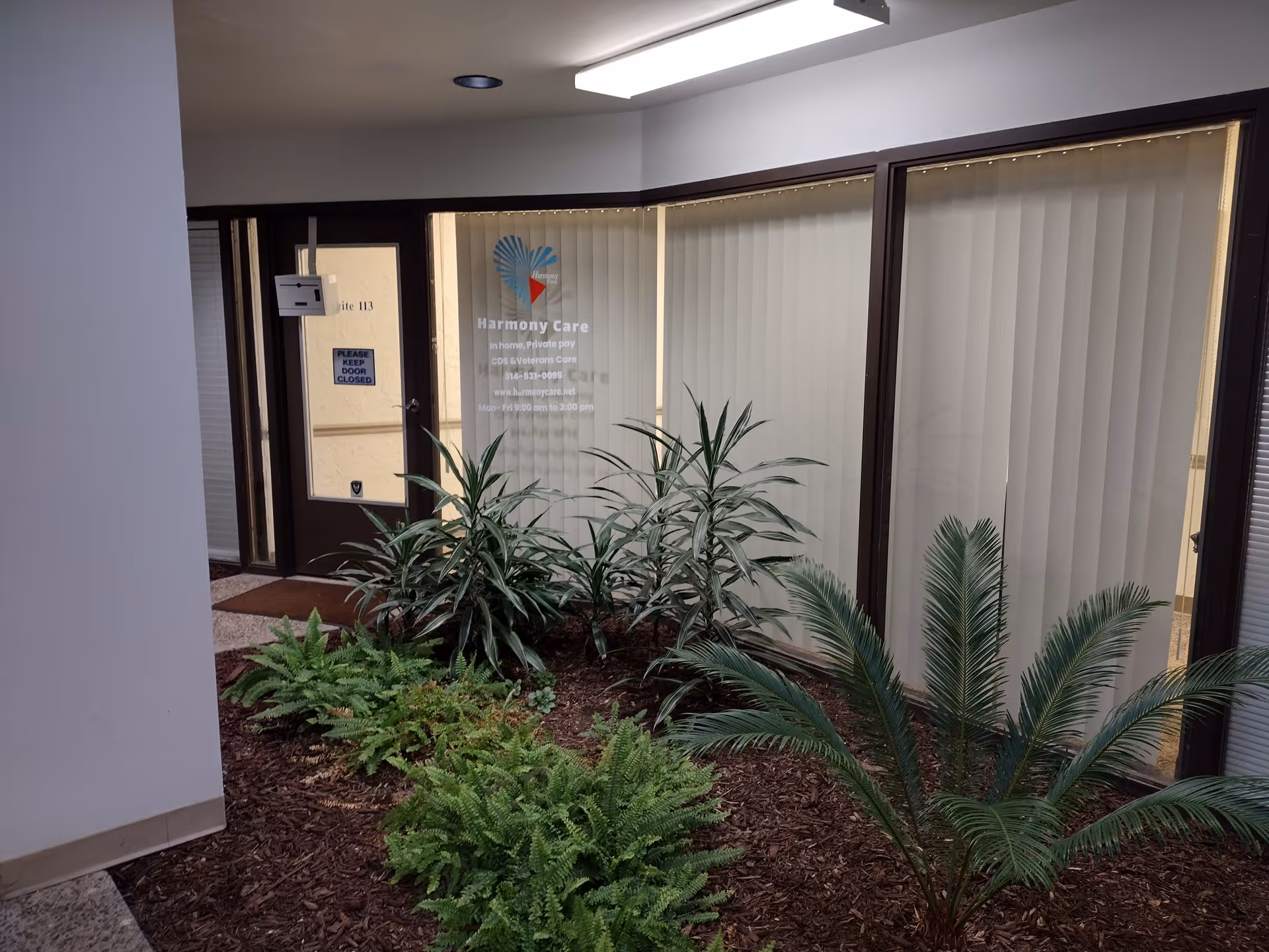 Exterior view of a Harmony Care entrance with glass doors, blinds, and surrounding plants.