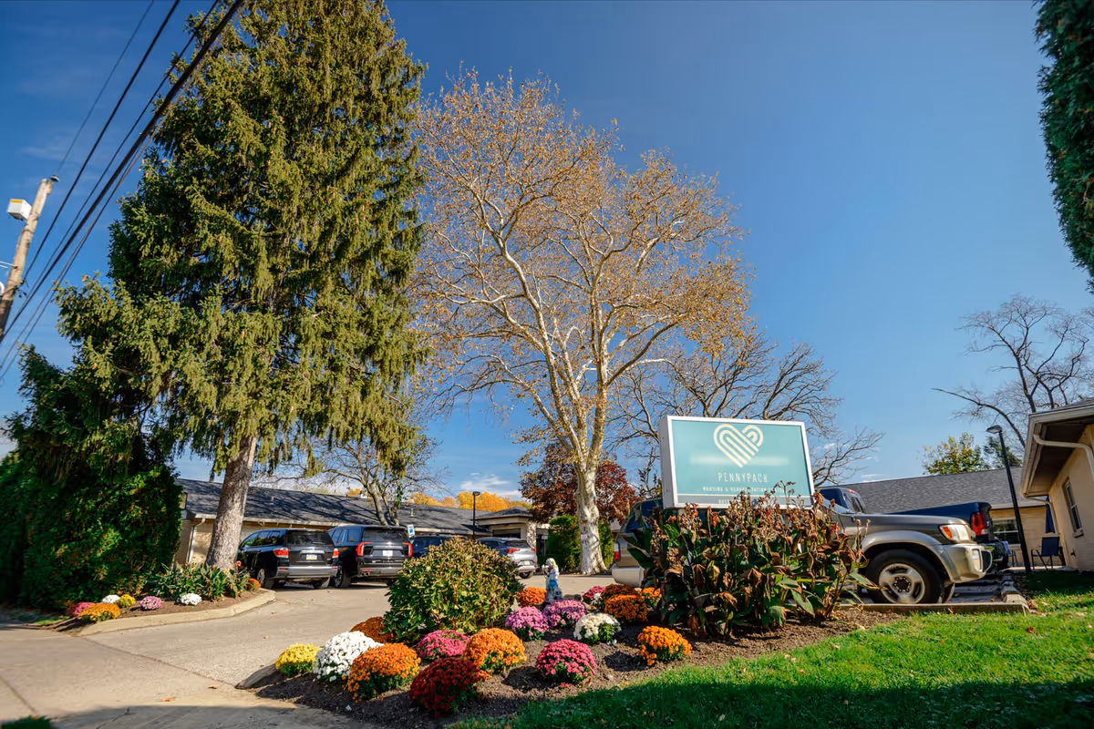 Exterior view of Pennypack Nursing and Rehabilitation Center with a landscaped garden featuring colorful flowers and bushes in the foreground, several parked cars, tall trees, and a clear blue sky.