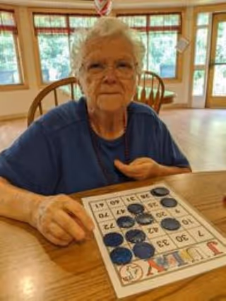 An elderly woman with white hair and glasses sitting at a wooden table playing a game of bingo. She is wearing a blue shirt and a beaded necklace. The room has large windows with a view of greenery outside and wooden chairs around the table.