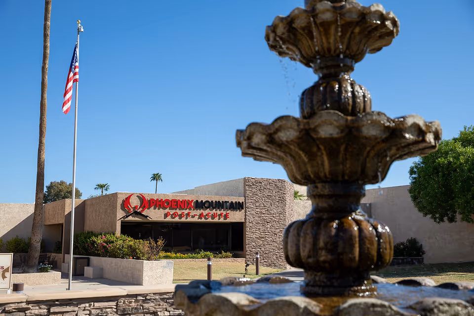 View of Phoenix Mountain Post-Acute building exterior with a stone façade, fountain, and American flag.